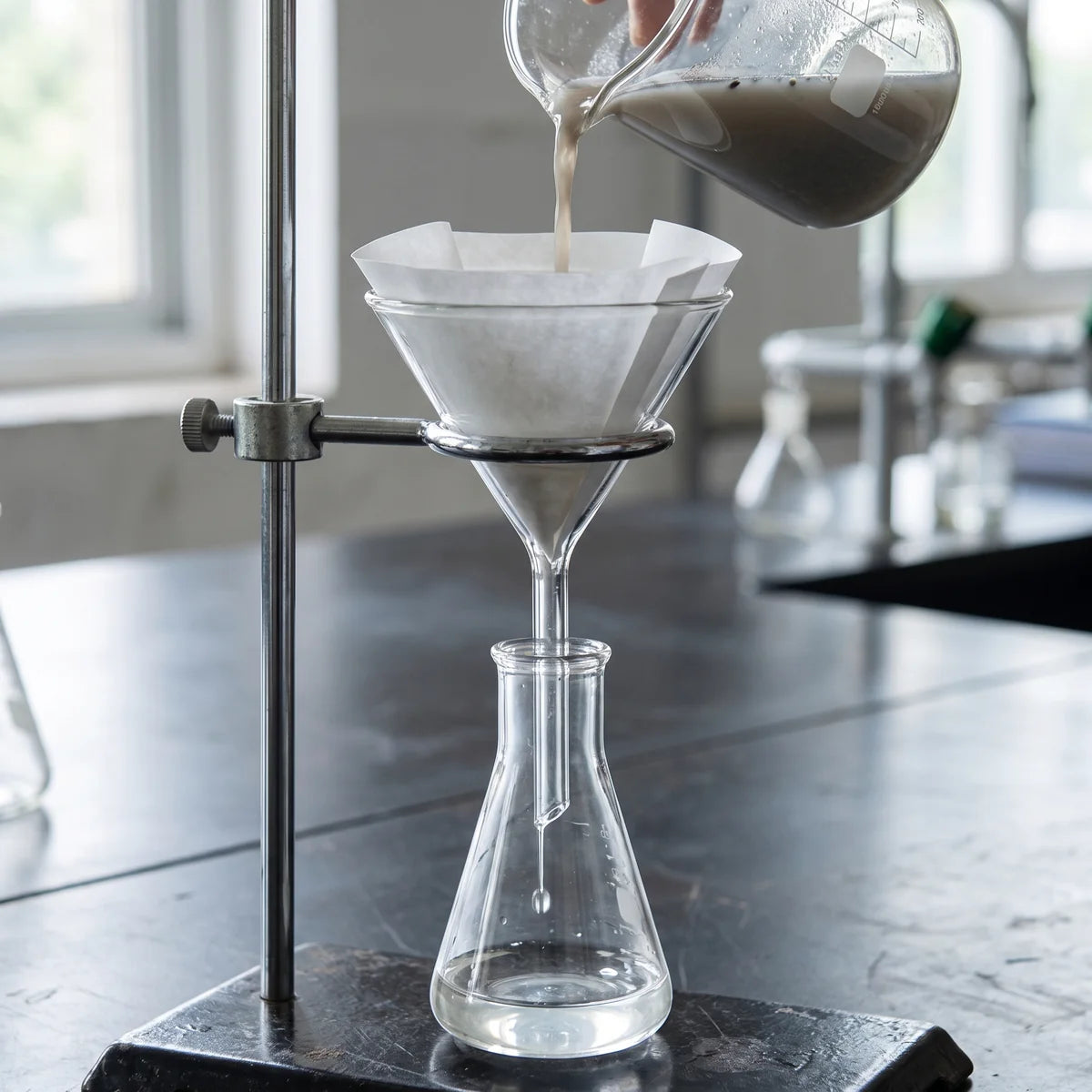 Cloudy mixture poured into a paper-lined glass filter funnel with clear filtrate dripping into a conical flask