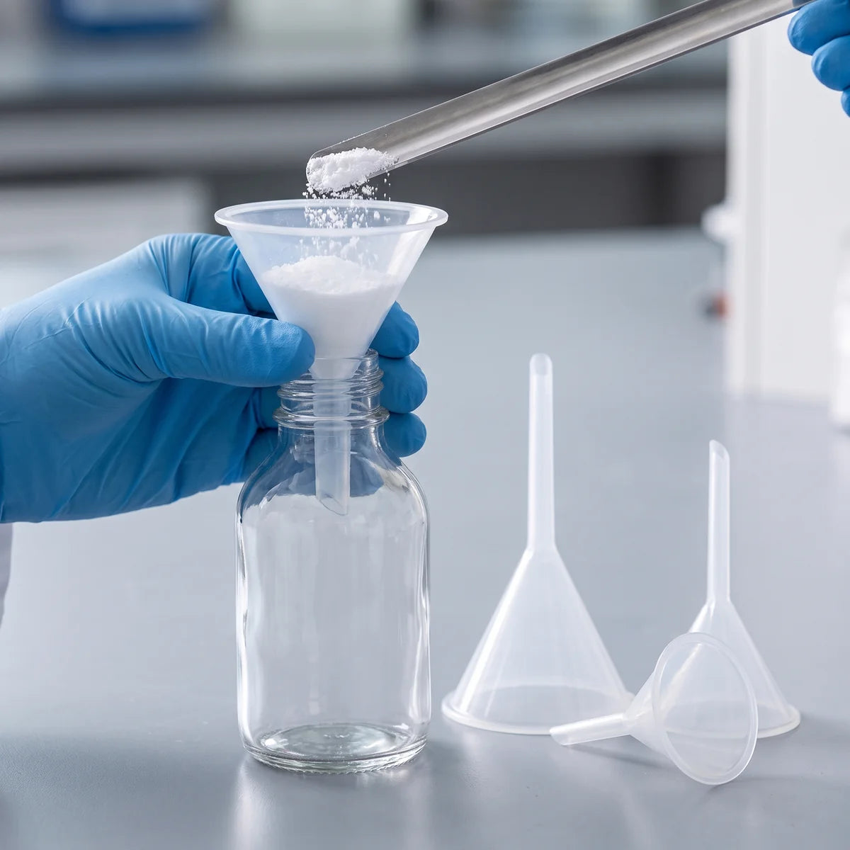Gloved hand holding a plastic powder funnel in a glass bottle while white powder is tipped in from a spatula