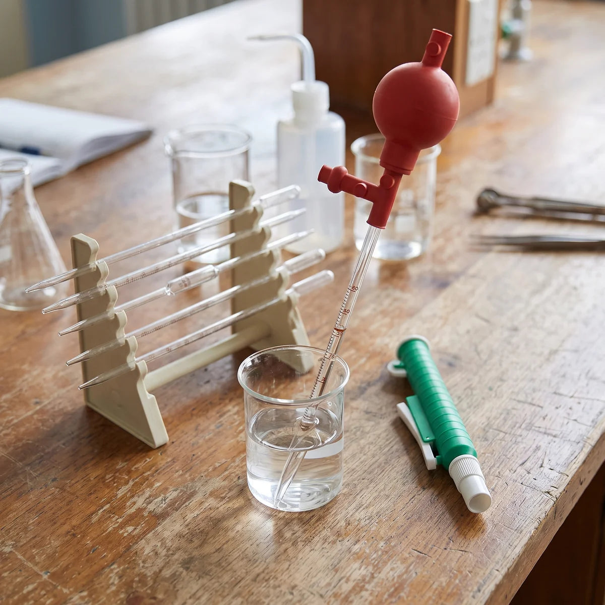 Red three-valve pipette filler bulb fitted to a graduated glass pipette in a beaker, with a pipette pump and pipette rack