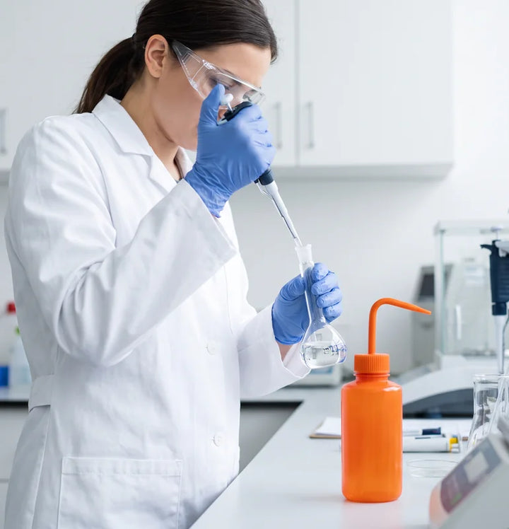 Lab technician in safety goggles and gloves beside an orange wash bottle