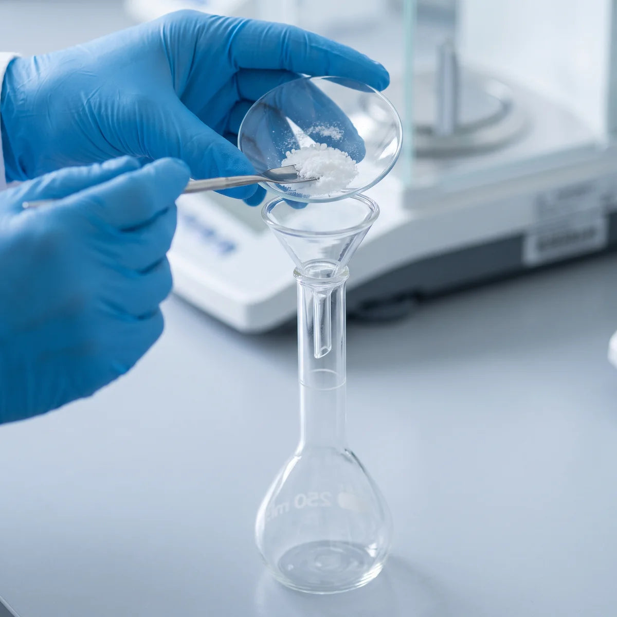 Gloved hands transferring weighed solid through a funnel into a volumetric flask beside a balance